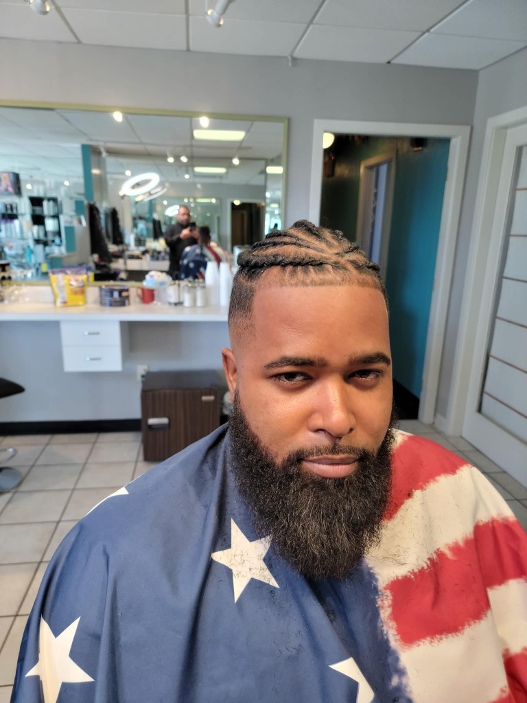 Client with cornrow braids and full beard in the chair, American flag barber cape